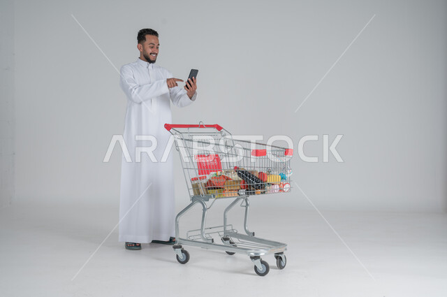 الاستمتاع بالتسوق في المولات والمراكز التجارية، شراء المستلزمات والحاجيات الضرورية، الاستفادة من الخصومات على المنتجات، بورتريه لرجل عربي خليجي سعودي يرتدي الثوب الابيض ويجر عربة التسوق أمامه يحمل بيده هاتف محمول، الجسم بالطول الكامل، خلفية بيضاء 
