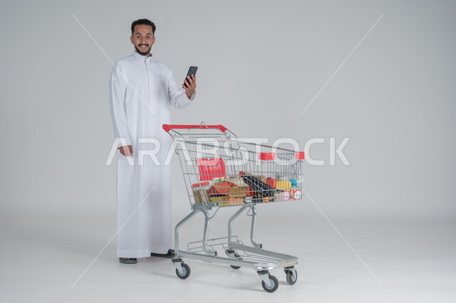 الاستمتاع بالتسوق في المولات والمراكز التجارية، شراء المستلزمات والحاجيات الضرورية، الاستفادة من الخصومات على المنتجات، بورتريه لرجل عربي خليجي سعودي يرتدي الثوب الابيض ويجر عربة التسوق أمامه يحمل بيده هاتف محمول، الجسم بالطول الكامل، خلفية بيضاء 
