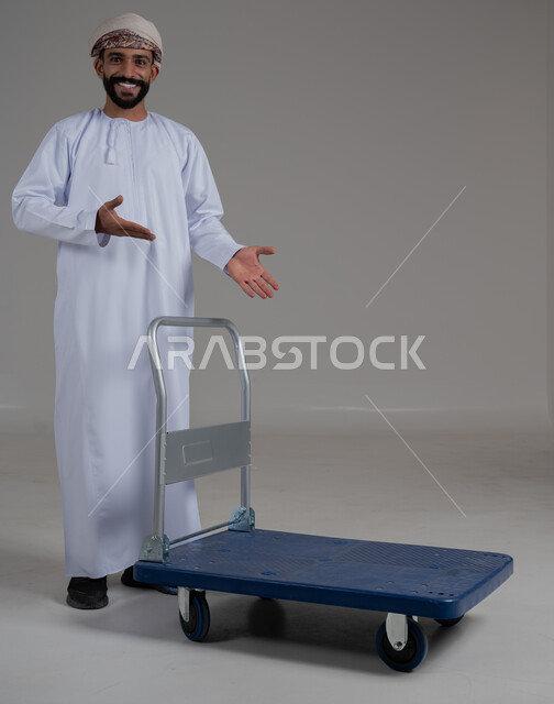تحديد موقع معين بإيماءات السرور والبشاشة، بورتريه لرجل عربي خليجي عماني يرتدي الدشداشة والعمامة يقوم بجر عربة دفع يدوية لنقل الأمتعة, الوقوف باستقامة والنظر إلى الكاميرا، اشارات وحركات يدين تدل على هذا المكان (هنا)، صورة الجسم بالطول الكامل، خلفية رمادية.