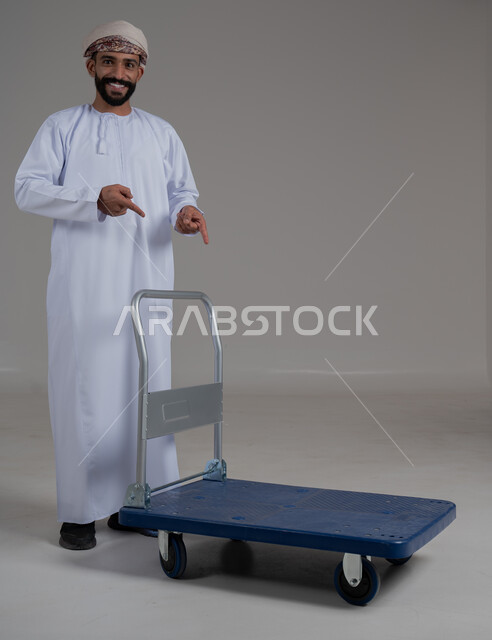 اشارات وحركات يدين تدل على هذا المكان (هنا)، تحديد موقع معين بإيماءات السرور والبشاشة، بورتريه لرجل عربي خليجي عماني يرتدي الدشداشة والعمامة يقوم بجر عربة دفع يدوية لنقل الأمتعة, الوقوف باستقامة والنظر إلى الكاميرا، صورة الجسم بالطول الكامل، خلفية رمادية.