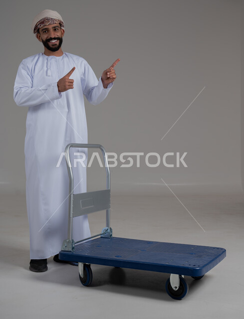 الوقوف باستقامة والنظر إلى الكاميرا، إشارات وحركات يدين تدل على هذا المكان (هنا)، تحديد موقع معين بإيماءات السرور والبشاشة، بورتريه لرجل عربي خليجي عماني يرتدي الدشداشة والعمامة يقوم بجر عربة دفع يدوية لنقل الأمتعة, صورة الجسم بالطول الكامل، خلفية رمادية.