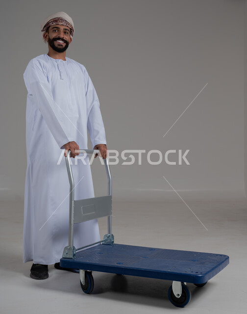 النظر الى الكاميرا بايماءات السعادة والسرور، خدمة توصيل الأمتعة الثقيلة الى الغرف في الفنادق، بورتريه لرجل عربي خليجي عماني يرتدي الدشداشة والعمامة يقوم بجر عربة دفع يدوية لنقل الأمتعة, الطول الكامل للجسم، خلفية رمادية