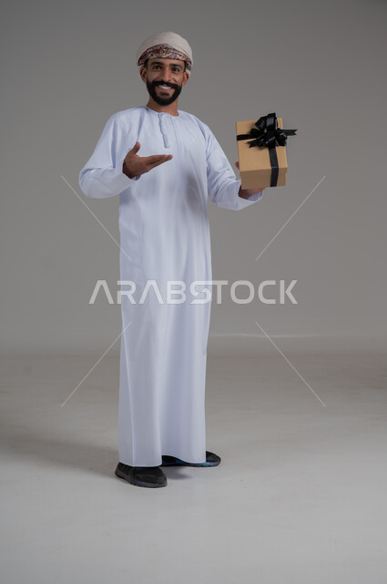 مفهوم الشكر والامتنان، تقديم الهدايا بالاعياد والمناسبات الخاصة، التعبير عن الحب والتقدير والاحترام، بورتريه لرجل عربي خليجي عماني يرتدي الدشداشة والعمامة يحمل صندوق هدية أصفر اللون وينظر إلى الكاميرا بايماءات الابتسامة، الجسم بالطول الكامل، خلفية بيضاء 