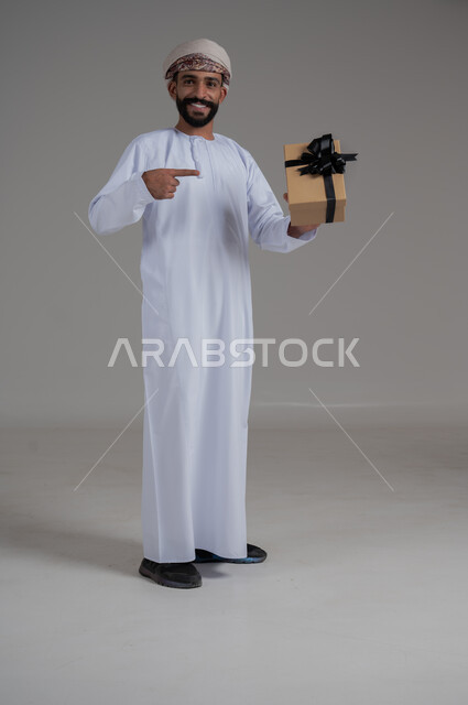 مفهوم الشكر والامتنان، تقديم الهدايا بالاعياد والمناسبات الخاصة، التعبير عن الحب والتقدير والاحترام، بورتريه لرجل عربي خليجي عماني يرتدي الدشداشة والعمامة يحمل صندوق هدية أصفر اللون وينظر إلى الكاميرا بايماءات الابتسامة، الجسم بالطول الكامل، خلفية بيضاء