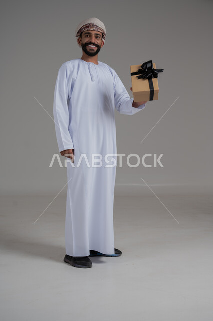 مفهوم الشكر والامتنان، تقديم الهدايا بالاعياد والمناسبات الخاصة، التعبير عن الحب والتقدير والاحترام، بورتريه لرجل عربي خليجي عماني يرتدي الدشداشة والعمامة يحمل صندوق هدية أصفر اللون وينظر إلى الكاميرا بايماءات الابتسامة، الجسم بالطول الكامل، خلفية بيضاء 