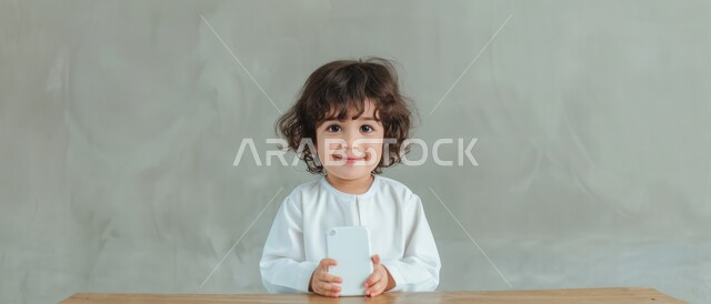 استعمال الأجهزة التقنية في دراسة المقررات المدرسية عن بعد, بورتريه لصبي عربي خليجي سعودي يرتدي الثوب التقليدي يجلس على الطاولة وينظر الى الكاميرا ويحمل الجوال, إيماءات الفرح والسرور , خلفية رمادية