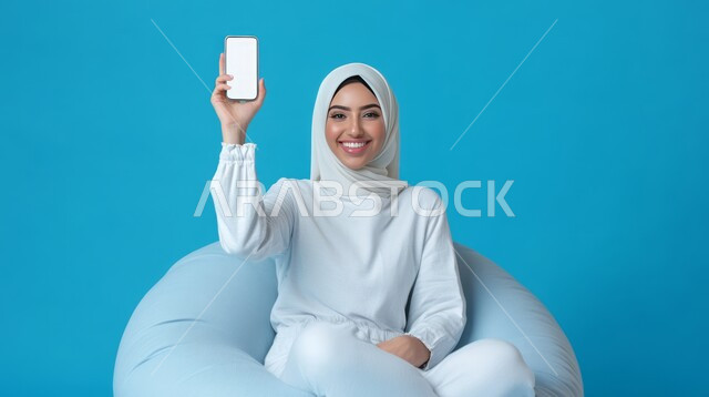 متاجر تسوق الكترونية, استخدام أجهزة التكنولوجيا الحديثة والمتطورة، بورتريه لامرأة عربية خليجية سعودية محجبة مبتسمة تجلس على الكرسي تحمل بيدها الهاتف المحمول, عرض شاشة بيضاء فارغة عبر الجوال، خلفية زرقاء