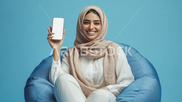 متاجر تسوق الكترونية, استخدام أجهزة التكنولوجيا الحديثة والمتطورة، بورتريه لامرأة عربية خليجية سعودية محجبة مبتسمة تجلس على الكرسي تحمل بيدها الهاتف المحمول, عرض شاشة بيضاء فارغة عبر الجوال، خلفية زرقاء