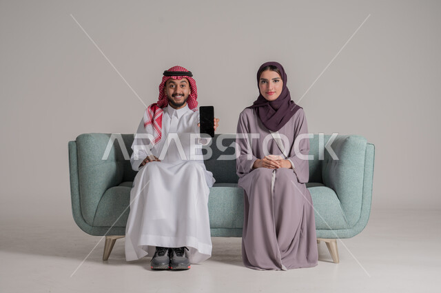Having fun on vacation, using modern technology devices, showing blank black screen on mobile phone, family love and affection, portrait of a Saudi Arabian Gulf couple wearing traditional clothes sitting on a comfortable sofa, full body length, gray background