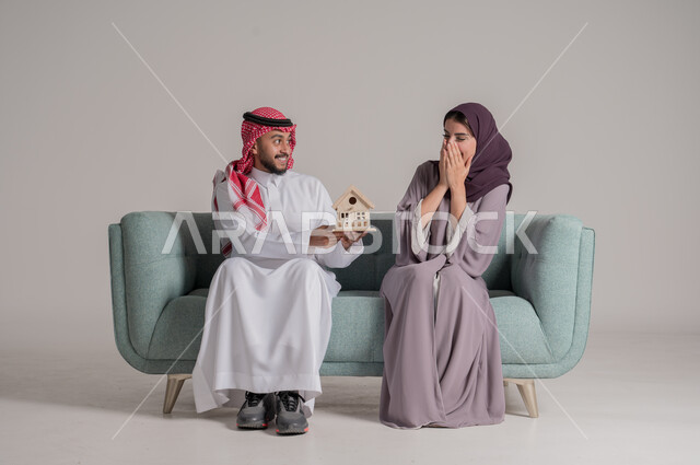 امتلاك سكن حديث والاستقرار العائلي، ايماءات السعادة والاندهاش، بناء أسرة ناجحة متفاهمة ومتماسكة، المحبة والمودة العائلية، بورتريه لرجل عربي خليجي سعودي يرتدي الثوب التقليدي والشماغ يحمل بيده مجسم صغير لبيت  ويفاجئ زوجته، الجسم بالطول الكامل، خلفية رمادية