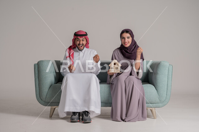 إمرأة تحمل بيدها مجسم بيت جديد، النظر الى الكاميرا بتعابير الفرح، امتلاك سكن حديث والاستقرار العائلي، مفهوم الاسكان والتمليك، ايماءات السعادة بشراء عقار جديد, بورتريه لزوجين عربيين خليجيين سعوديين يرتديان الملابس التقليدية يجلسان على كنبه مريحة، خلفية رمادية