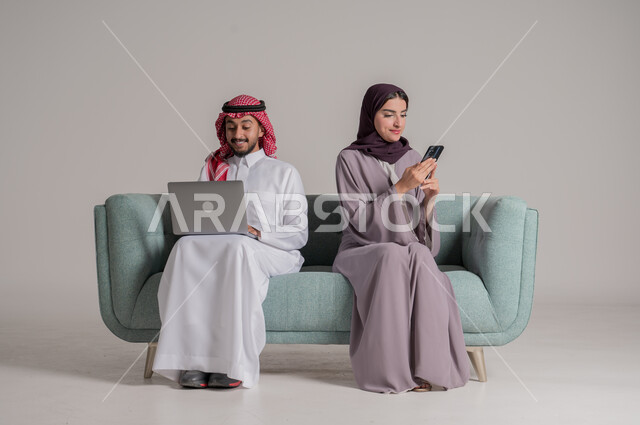 Facilitating the buying and selling process through electronic applications, the concept of online shopping and ordering products online, a portrait of a Saudi Arabian Gulf couple wearing traditional clothes sitting on a comfortable sofa and working on modern technical devices, full body length, and a gray background