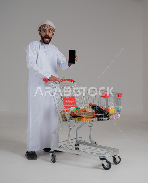 الحماس للاستفادة من الخصومات على المنتجات، الاستمتاع بالتسوق في المولات والمراكز التجارية، شراء المستلزمات والحاجيات الضرورية، بورتريه لرجل عربي خليجي عماني يرتدي الدشداشة والعمامة يجر أمامه عربة مشتريات ويحمل بيده هاتف محمول، الجسم بالطول الكامل، خلفية بيضاء 