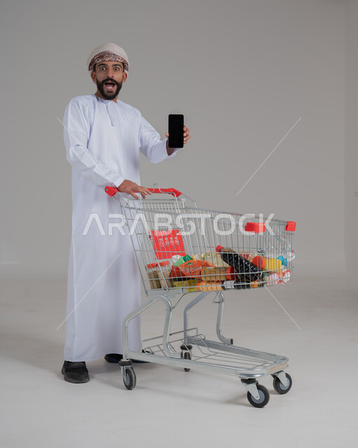 Excitement to benefit from discounts on products, enjoy shopping in malls and shopping centers, buy necessary supplies and necessities, portrait of an Arab Gulf Omani man wearing a dishdasha and turban pulling a shopping cart in front of him and holding a mobile phone in his hand, full body, white background