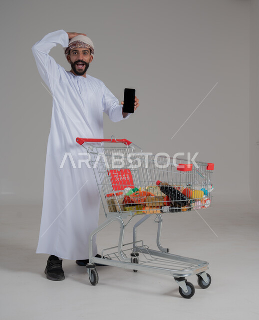 الحماس للاستفادة من الخصومات على المنتجات، الاستمتاع بالتسوق في المولات والمراكز التجارية، شراء المستلزمات والحاجيات الضرورية، بورتريه لرجل عربي خليجي عماني يرتدي الدشداشة والعمامة يجر أمامه عربة مشتريات ويحمل بيده هاتف محمول، الجسم بالطول الكامل، خلفية بيضاء 