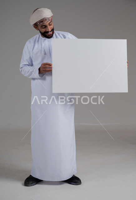 إعلانات وعروض تسويقية، استخدام لوحات توضيحية ووسائل تعليمية، بورتريه لرجل عربي خليجي عماني مبتسم يرتدي الدشداشه والعمامة  يحمل بيده لوح ابيض كبير (موك اب فارغ)، ايماءات السعادة والسرور، الطول الكامل للجسم، خلفية رماديه