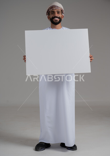 استخدام لوحات توضيحية ووسائل تعليمية، بورتريه لرجل عربي خليجي عماني مبتسم يرتدي الدشداشه والعمامة  يحمل بيده لوح ابيض كبير (موك اب فارغ)، النظر الى الكاميرا بايماءات السعادة والسرور، إعلانات وعروض تسويقية، الطول الكامل للجسم، خلفية رماديه