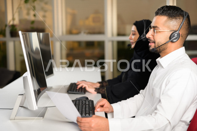 A Saudi Arabian Gulf employee working in customer service and holding papers in his hand, an employee and an employee wearing a headset, making inquiries and inquiries, answering customers’ questions, making a video call through a computer, following up a