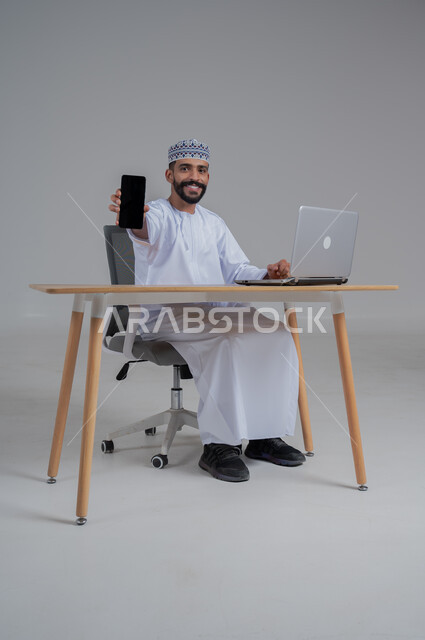 مهن ووظائف مكتبيه عمانية، استخدام التقنيات الحديثة والمتطورة، بورتريه لرجل عربي خليجي عماني  مبتسم ترتدي الدشداشة والعمامة يجلس على الكرسي أمام الحاسوب المحمول يحمل بيده الجوال بشاشة سوداء فارغه، النظر الى الكاميرا بايماءات السعادة والسرور، خلفية بيضاء