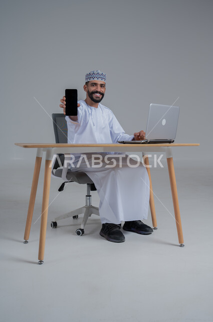 مهن ووظائف مكتبيه عمانية، استخدام التقنيات الحديثة والمتطورة، بورتريه لرجل عربي خليجي عماني  مبتسم ترتدي الدشداشة والعمامة يجلس على الكرسي أمام الحاسوب المحمول يحمل بيده الجوال بشاشة سوداء فارغه، النظر الى الكاميرا بايماءات السعادة والسرور، خلفية بيضاء 