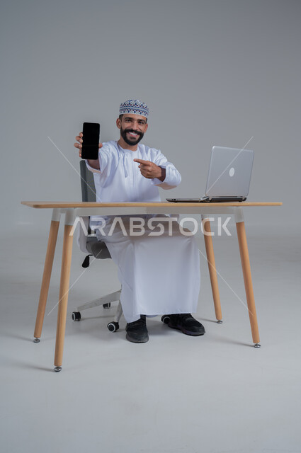 مهن ووظائف مكتبيه عمانية، استخدام التقنيات الحديثة والمتطورة، بورتريه لرجل عربي خليجي عماني  مبتسم ترتدي الدشداشة والعمامة يجلس على الكرسي أمام الحاسوب المحمول يحمل بيده الجوال بشاشة سوداء فارغه، النظر الى الكاميرا بايماءات السعادة والسرور، خلفية بيضاء 