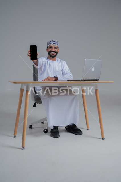 مهن ووظائف مكتبيه عمانية، استخدام التقنيات الحديثة والمتطورة، بورتريه لرجل عربي خليجي عماني  مبتسم ترتدي الدشداشة والعمامة يجلس على الكرسي أمام الحاسوب المحمول يحمل بيده الجوال بشاشة سوداء فارغه، النظر الى الكاميرا بايماءات السعادة والسرور، خلفية بيضاء 