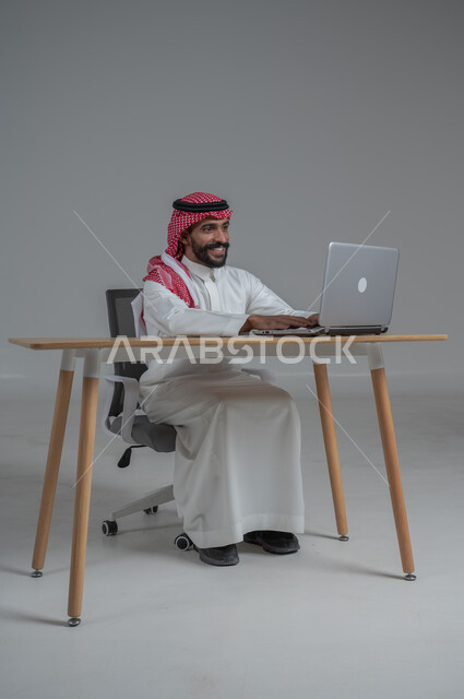 مهن ووظائف مكتبيه سعودية، استخدام التقنيات الحديثة والمتطورة، بورتريه لرجل عربي خليجي عماني  مبتسم يرتدي الثوب الابيض والشماغ يجلس على الكرسي أمام الحاسوب المحمول، النظر بايماءات السعادة والسرور، خلفية بيضاء 