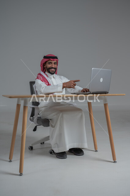 مهن ووظائف مكتبيه سعودية، استخدام التقنيات الحديثة والمتطورة، بورتريه لرجل عربي خليجي عماني  مبتسم يرتدي الثوب الابيض والشماغ يجلس على الكرسي أمام الحاسوب المحمول، النظر بايماءات السعادة والسرور، خلفية بيضاء 