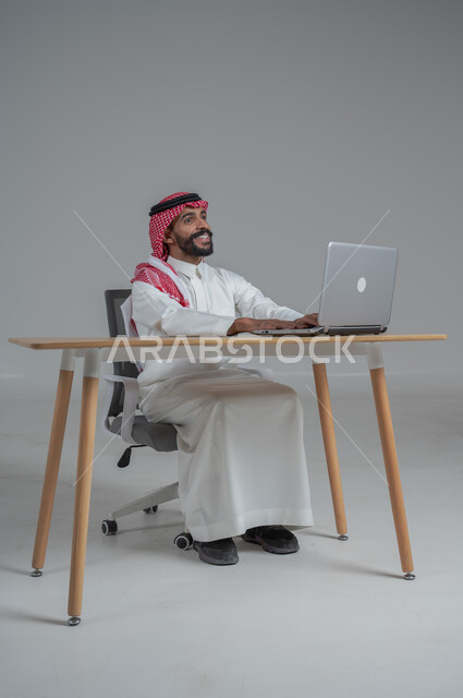 مهن ووظائف مكتبيه سعودية، استخدام التقنيات الحديثة والمتطورة، بورتريه لرجل عربي خليجي عماني  مبتسم يرتدي الثوب الابيض والشماغ يجلس على الكرسي أمام الحاسوب المحمول، النظر بايماءات السعادة والسرور، خلفية بيضاء 