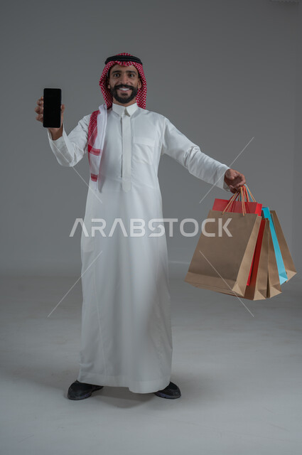 الحماس للاستفادة من الخصومات على المنتجات، الاستمتاع بالتسوق في المولات والمراكز التجارية، شراء المستلزمات والحاجيات الضرورية، بورتريه لرجل عربي خليجي سعودي يرتدي الثوب الابيض والشماغ يقوم بعرض شاشة سوداء فارغة، الجسم بالطول الكامل، خلفية بيضاء