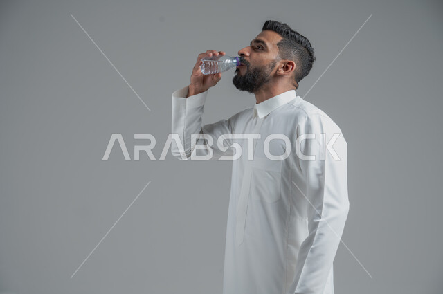 أهمية شرب الماء النقي للمحافظة على بشرة نضرة، بورتريه لرجل عربي خليجي سعودي يرتدي الثوب التقليدي ويشرب المياه من عبوة بلاستيكية، المحافظة على سلامة الجهاز المناعي، التخلص من سموم الجسم، الحفاظ على صحة الكلى ومعالجتها، الجسم بالطول الكامل، خلفية رمادية