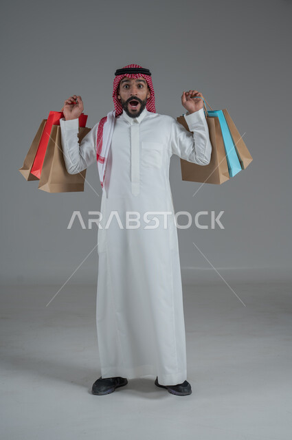 الحماس للاستفادة من الخصومات على المنتجات، الاستمتاع بالتسوق في المولات والمراكز التجارية، شراء المستلزمات والحاجيات الضرورية، بورتريه لرجل عربي خليجي سعودي يرتدي الثوب الابيض والشماغ يحمل بيده أكياس ورقية ملونة، الجسم بالطول الكامل، خلفية بيضاء 