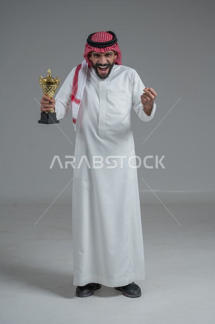 Enthusiasm and interaction with the team, gestures of passion and encouragement for the team, watching and following a football match, portrait of a smiling Saudi Arabian Gulf man wearing a traditional thobe and shemagh holding the World Cup in his hand, different sports clubs, gray background