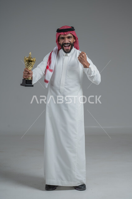 الحماس والتفاعل مع المنتخب، إيماءات الشغف والتشجيع للفريق، مشاهدة ومتابعة مباراة كرة القدم، بورتريه لرجل عربي خليجي سعودي مبتسم يرتدي الثوب التقليدي والشماغ يحمل بيده كأس العالم، أنديه رياضية مختلفة، خلفية رمادية