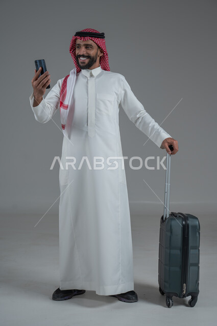 Expressions of joy and readiness for a fun summer trip, concept of tourism in summer vacation, taking recreational tours, portrait of a Saudi Arabian Gulf man wearing traditional thobe and shemagh holding a mobile phone and luggage bag, full body, white background