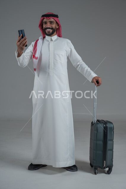 Expressions of joy and readiness for a fun summer trip, concept of tourism in summer vacation, taking recreational tours, portrait of a Saudi Arabian Gulf man wearing traditional thobe and shemagh holding a mobile phone and luggage bag, full body, white background