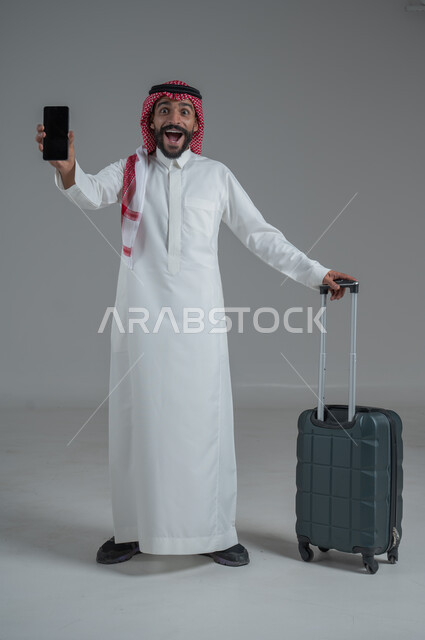 Expressions of joy and readiness for a fun summer trip, concept of tourism in summer vacation, taking recreational tours, portrait of a Saudi Arabian Gulf man wearing traditional thobe and shemagh holding a mobile phone and luggage bag, full body, white background