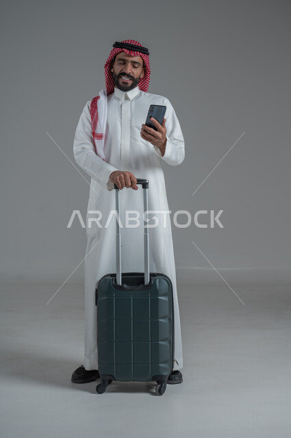 تعابير الفرح والاستعداد لقضاء رحلة صيفية ممتعة، مفهوم السياحة في إجازة الصيف، القيام بالجولات الترفيهية، بورتريه لرجل عربي خليجي سعودي يرتدي الثوب التقليدي والشماغ يحمل بيده الهاتف المحمول وحقيبة الأمتعة، الجسم بالطول الكامل، خلفية بيضاء 