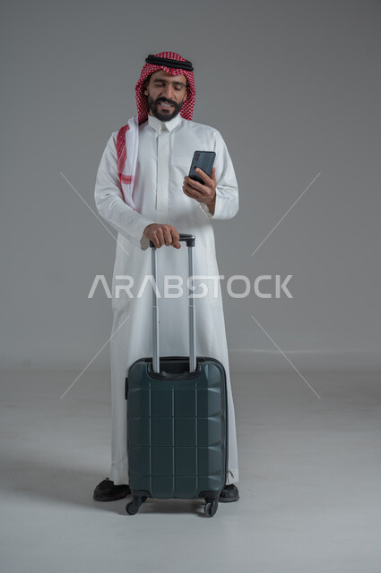 تعابير الفرح والاستعداد لقضاء رحلة صيفية ممتعة، مفهوم السياحة في إجازة الصيف، القيام بالجولات الترفيهية، بورتريه لرجل عربي خليجي سعودي يرتدي الثوب التقليدي والشماغ يحمل بيده الهاتف المحمول وحقيبة الأمتعة، الجسم بالطول الكامل، خلفية بيضاء 