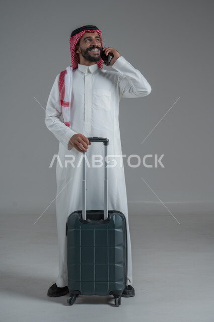 Expressions of joy and readiness for a fun summer trip, concept of tourism in summer vacation, taking recreational tours, portrait of a Saudi Arabian Gulf man wearing traditional thobe and shemagh holding a mobile phone and luggage bag, full body, white background