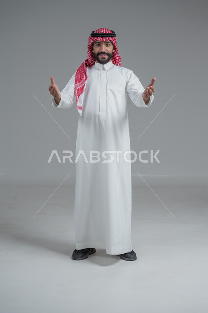 مفهوم الرجولة والاهتمام بالمظهر الخارجي، الثقة بالنفس والجدية، الوقوف باستقامة وثبات، بورتريه لرجل عربي خليجي سعودي يرتدي الشماغ والثوب التقليدي ينظر الى الكاميرا بتعابير السرور،الطول الكامل للجسم، خلفية رمادية