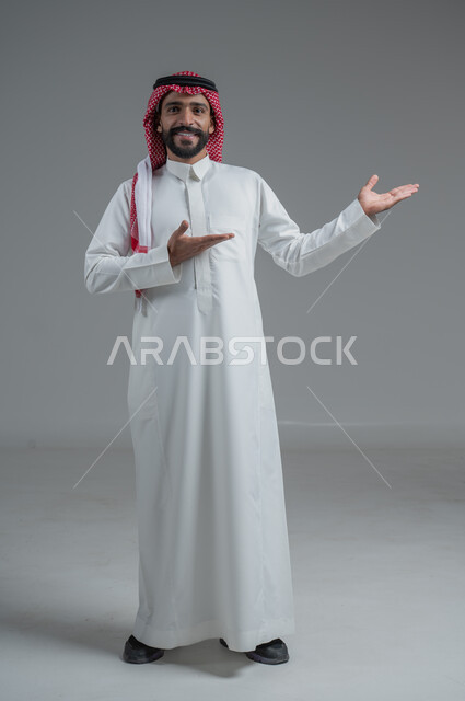 The concept of masculinity and concern for external appearance, self-confidence and seriousness, standing straight and steadfast, portrait of a Saudi Arabian Gulf man wearing a traditional shemagh and thobe looking at the camera with an expression of pleasure, full body length, gray background