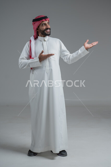 مفهوم الرجولة والاهتمام بالمظهر الخارجي، الثقة بالنفس والجدية، الوقوف باستقامة وثبات، بورتريه لرجل عربي خليجي سعودي يرتدي الشماغ والثوب التقليدي ينظر الى الكاميرا بتعابير السرور،الطول الكامل للجسم، خلفية رمادية