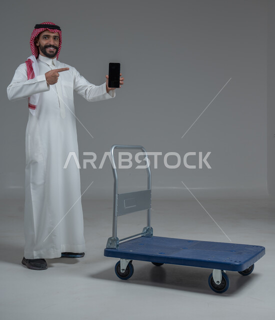 الاستمتاع بالتسوق في المولات والمراكز التجارية، عرض شاشة هاتف محمول سوداء فارغة، الاستفادة من الخصومات والتنزيلات، بورتريه لرجل عربي خليجي سعودي يرتدي الثوب التقليدي و الشماغ يقف ويمسك الجوال بيده امام عربة للمشتريات الثقيلة، الجسم بالطول الكامل، خلفية بيضاء