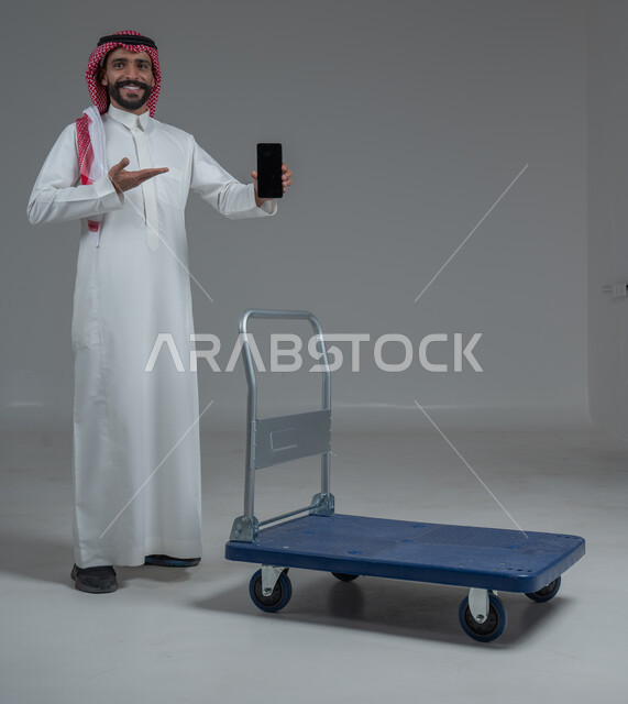 الاستمتاع بالتسوق في المولات والمراكز التجارية، عرض شاشة هاتف محمول سوداء فارغة، الاستفادة من الخصومات والتنزيلات، بورتريه لرجل عربي خليجي سعودي يرتدي الثوب التقليدي و الشماغ يقف ويمسك الجوال بيده امام عربة للمشتريات الثقيلة، الجسم بالطول الكامل، خلفية بيضاء