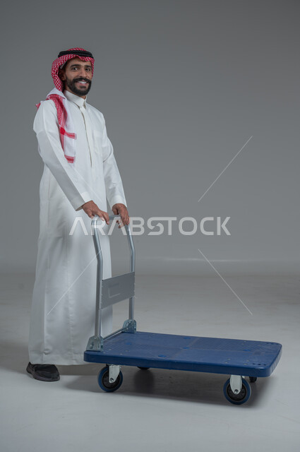 النظر الى الكاميرا بايماءات السعادة والسرور، خدمة توصيل الأمتعة الثقيلة الى الغرف في الفنادق، بورتريه لرجل عربي خليجي سعودي يرتدي الثوب التقليدي والشماغ يقوم بجر عربة دفع يدوية لنقل الأمتعة, الطول الكامل للجسم، خلفية رمادية