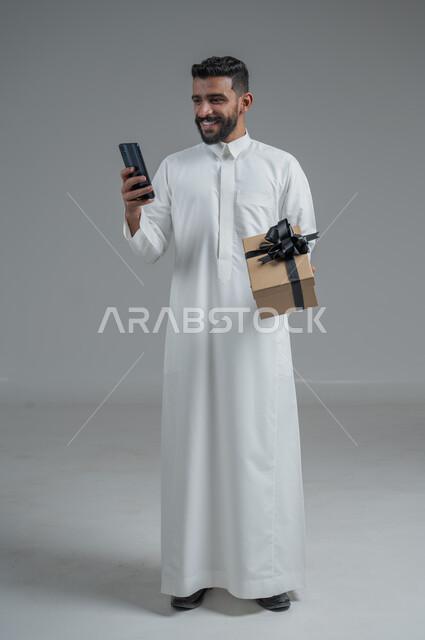 مفهوم الشكر والامتنان، تقديم الهدايا بالاعياد والمناسبات الخاصة، التعبير عن الحب والتقدير والاحترام، بورتريه لرجل عربي خليجي سعودي يرتدي ثوب أبيض يحمل صندوق هدية أسود اللون يحمل بيده الهاتف المحمول بايماءات السعادة والسرور، خلفية رماية