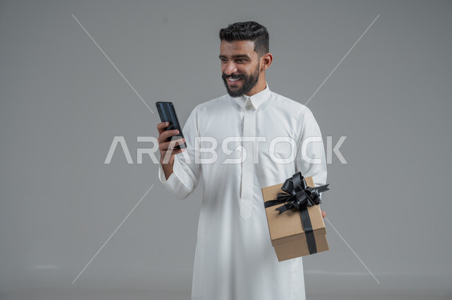 مفهوم الشكر والامتنان، تقديم الهدايا بالاعياد والمناسبات الخاصة، التعبير عن الحب والتقدير والاحترام، بورتريه لرجل عربي خليجي سعودي يرتدي ثوب أبيض يحمل صندوق هدية أسود اللون يحمل بيده الهاتف المحمول بايماءات السعادة والسرور، خلفية رماية