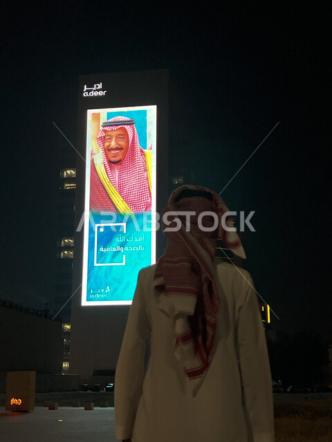 الاحتفال بالأعياد والمناسبات الوطنية، صورة من الخلف لرجل عربي سعودي خليجي يرتدي الثوب التقليدي مع الشماغ ينظر إلى لوحة طرقية لصاحب السمو الملكي السعودي، صورة الملك سلمان بن عبدالعزيز آل سعود، ذكرى اليوم الوطني السعودي94، معالم المملكة العربية السعودية المشهورة