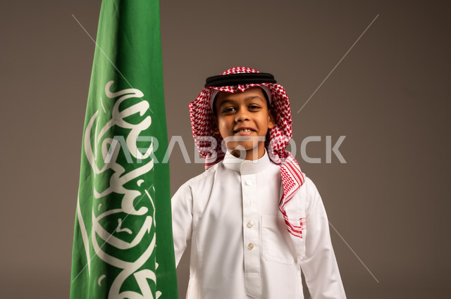 Saudi National Day 23 September, pride and honor in having a national identity, love of the homeland and belonging to it, close-up portrait of a Saudi Arabian Gulf boy wearing traditional dress and shemagh holding the Kingdom's flag in his hand and looking at the camera, celebrating Flag Day 11 March, gray background